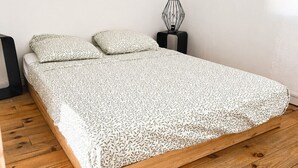 Sängyssä on kuviollinen päiväpeite, kaksi tyynyä, geometrisesti muotoiltu lamppu yöpöydällä ja puulattia.