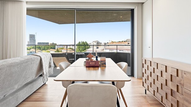 Ein modernes Wohnzimmer mit einer Couch, einem Esstisch und Stühlen. Ein großes Fenster bietet Blick auf Gebäude und Bäume.