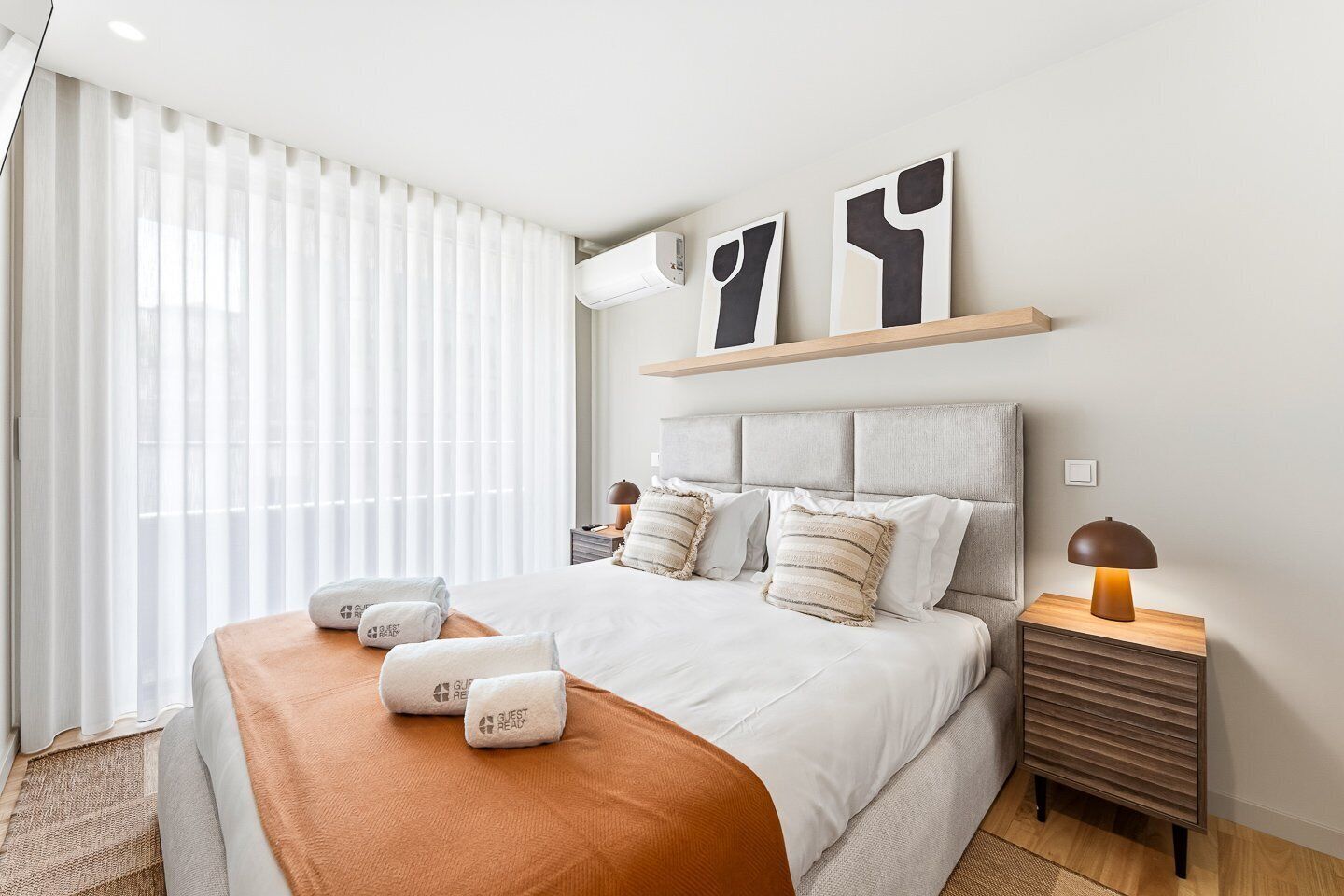 A modern hotel room with a large bed, a bedside table with a lamp, a window with sheer curtains, and abstract wall art.