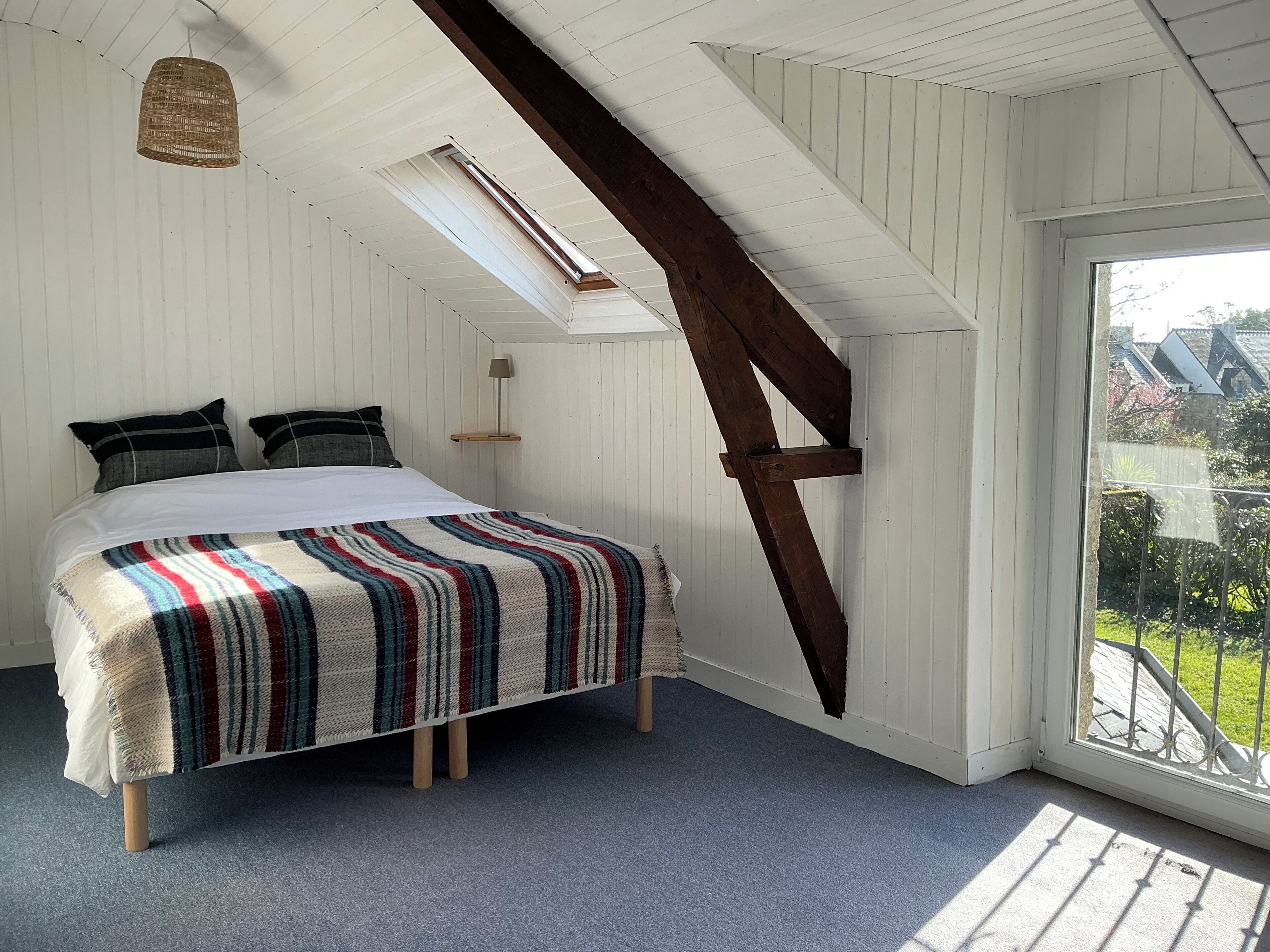 A bedroom with a sloped ceiling, a bed with a striped blanket, a hanging light fixture, and a window with a view of greenery.