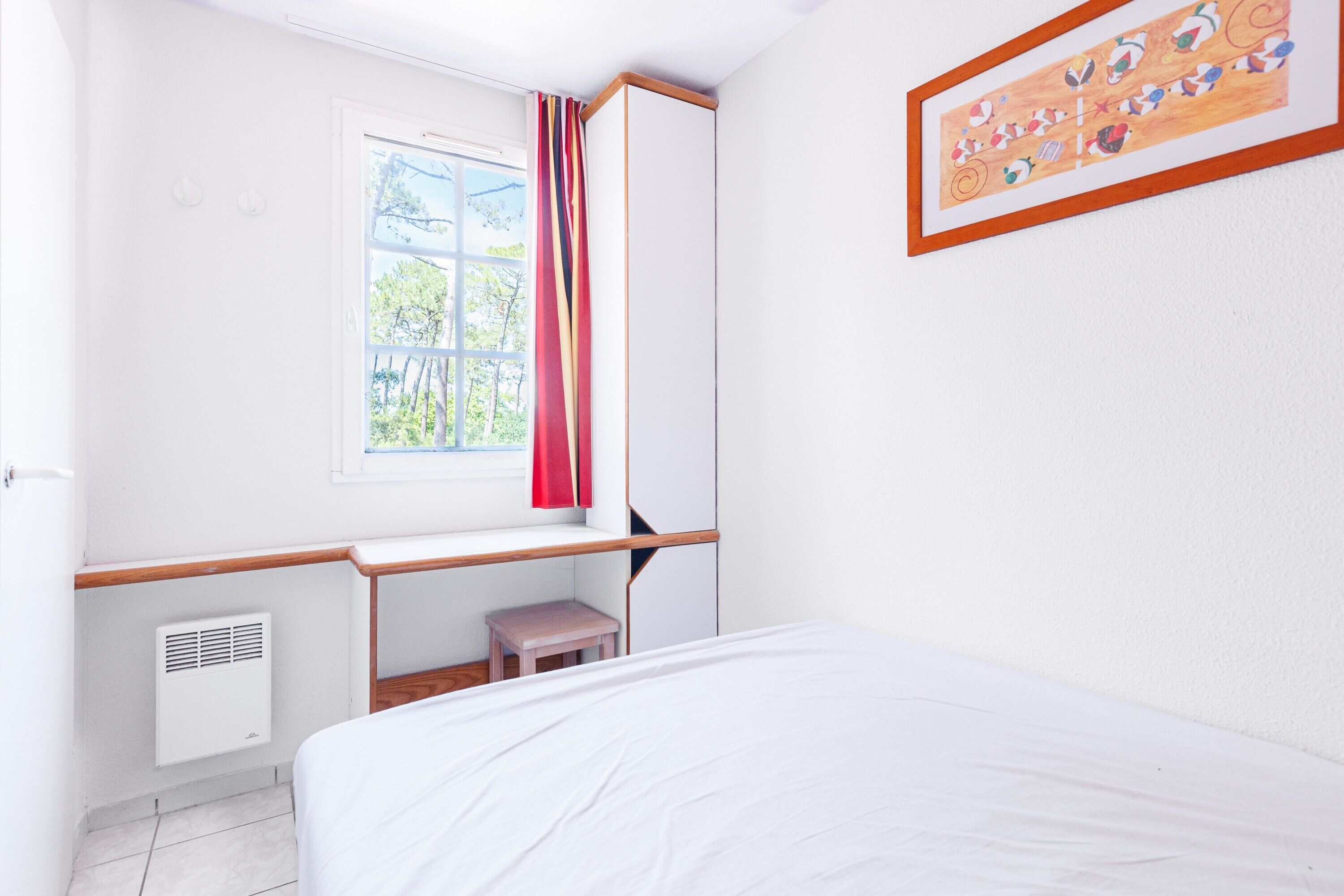 Una stanza con un letto, una finestra con tende, un termosifone, un tavolino e un quadro appeso al muro.