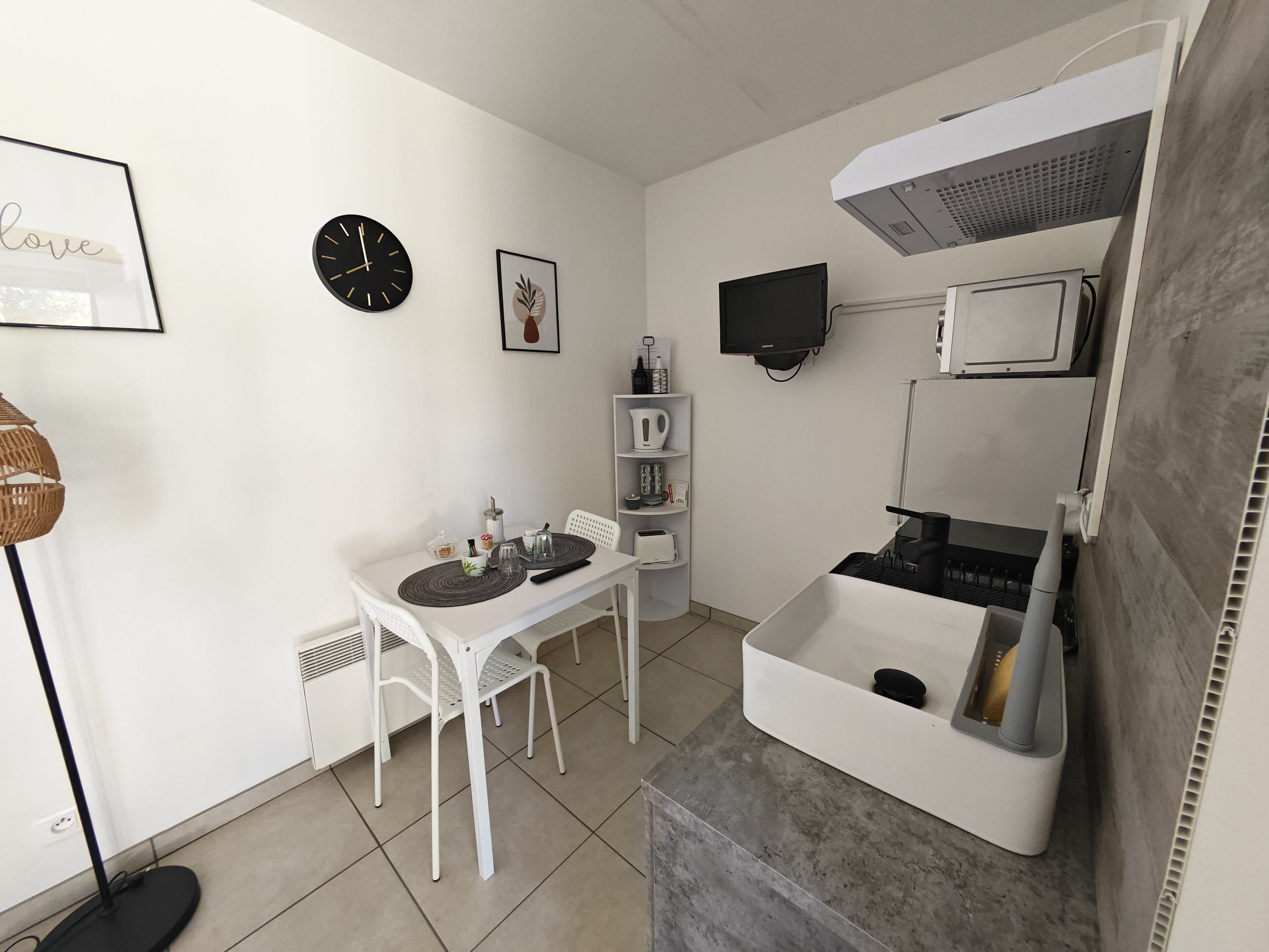 A compact kitchen with a dining table, chairs, a clock, a framed artwork, a television, and a built-in microwave.