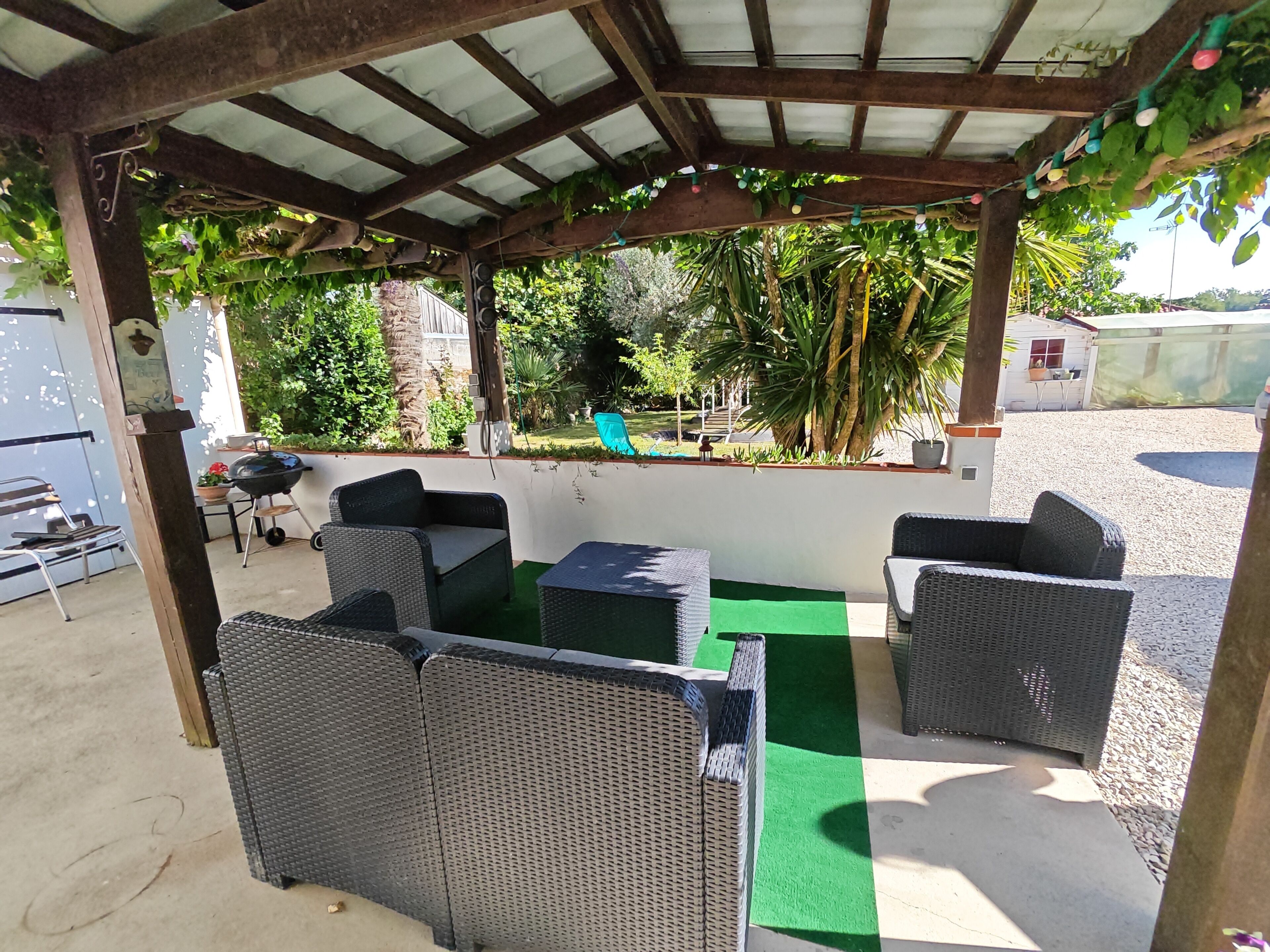 Un espacio exterior techado con mobiliario de mimbre, un tapete verde y vistas a un jardín con palmeras y una barbacoa.