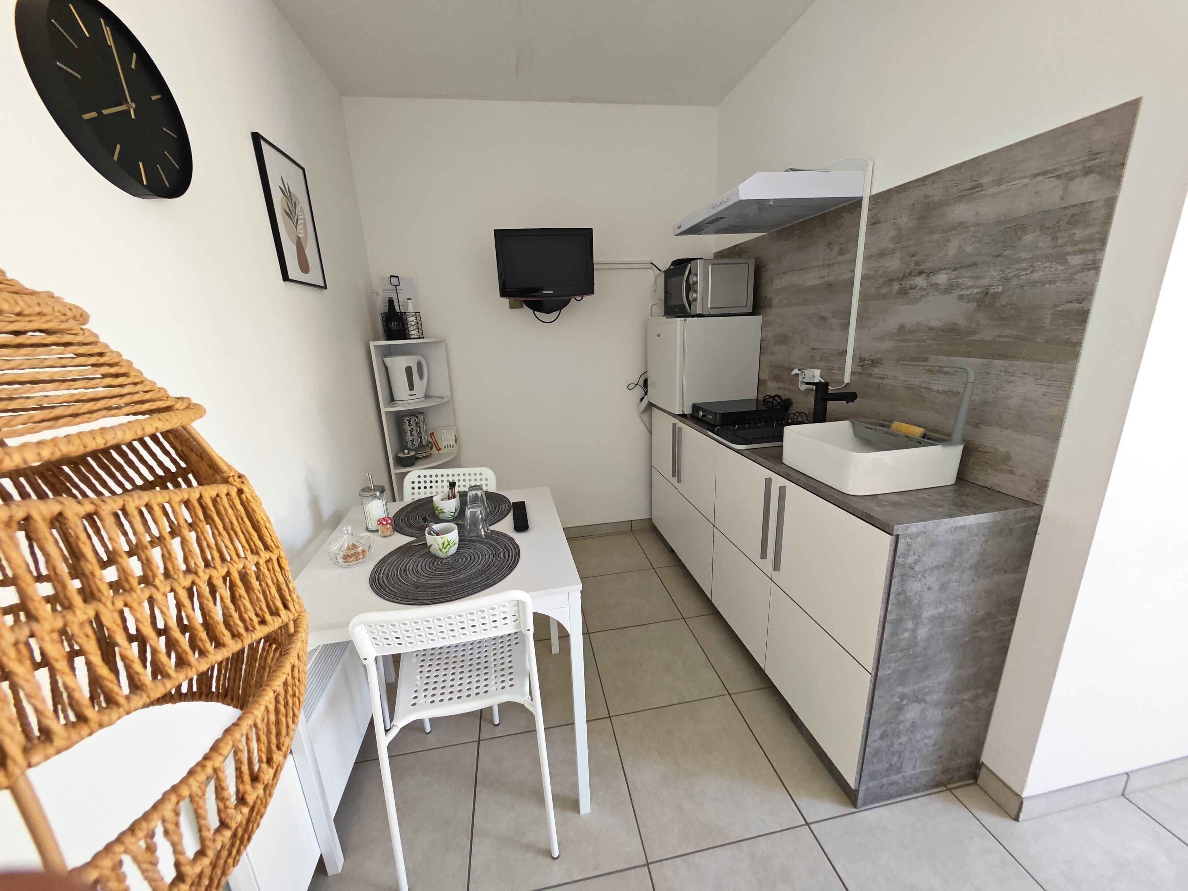 A compact kitchen with a microwave, stove, and sink. There is a dining table with chairs and a wicker chair. A clock and framed picture are on the wall.