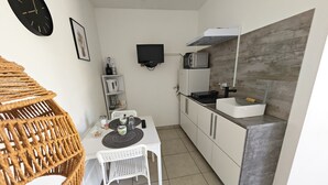 Een kleine keuken met een magnetron, fornuis en wastafel. Er is een eettafel met stoelen, en een rieten fauteuil. Een klok en een lijst met foto hangen aan de muur.
