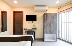 Ein Hotelzimmer mit einem Bett, einem Schreibtisch mit Stuhl, einem an der Wand befestigten Fernseher, einer Klimaanlage und einem großen Fenster mit Blick.