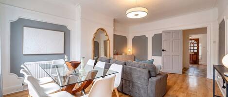 A modern living room with a glass dining table, white chairs, a grey sofa, and a wooden floor.