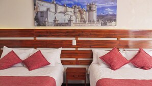 A hotel room with two beds, each with white linens and red pillows, a wooden headboard, a small wooden nightstand, and a painting of a cityscape on the wall.