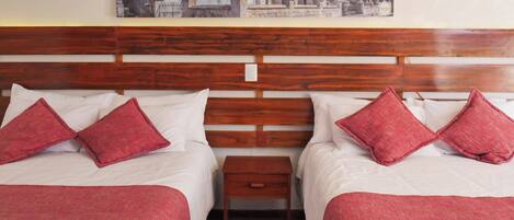 A hotel room with two beds, each with white linens and red pillows, a wooden headboard, a small wooden nightstand, and a painting of a cityscape on the wall.