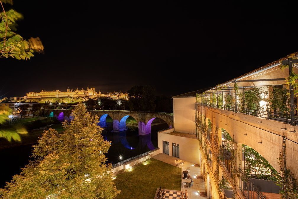 Vista nocturna de un edificio a la orilla del río con arcos iluminados, un puente y una zona exterior bien iluminada.