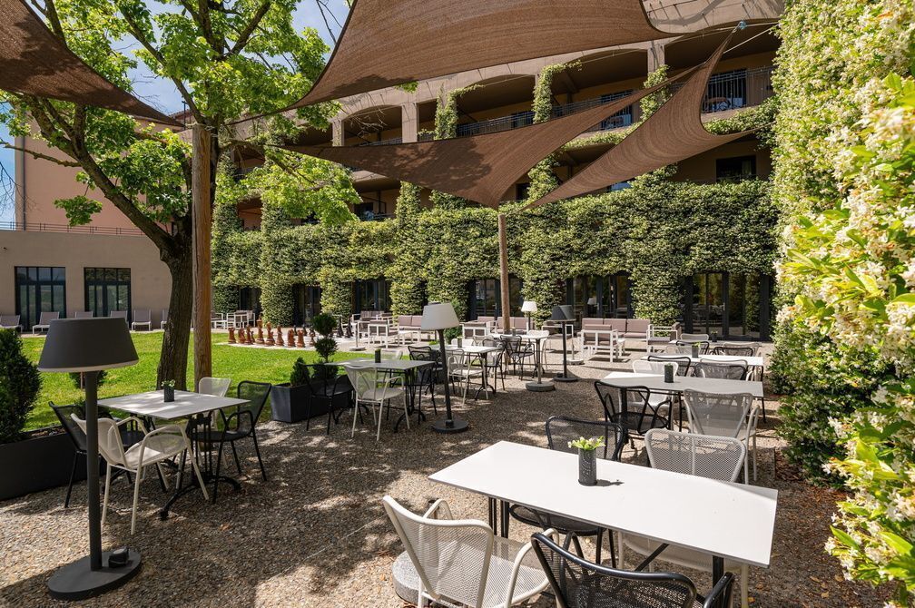 Un espace café en plein air, avec des tables et des chaises, entouré de verdure et bordé d’un immeuble doté de balcons.