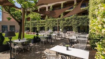 Un espace café en plein air, avec des tables et des chaises, entouré de verdure et bordé d’un immeuble doté de balcons.