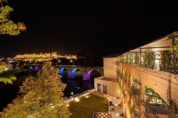 A nighttime view of a riverside building with illuminated arches, a bridge, and a well-lit outdoor area.