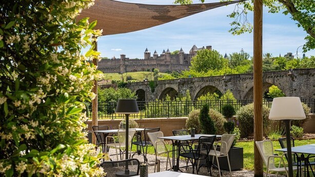 An outdoor patio with tables and chairs, a wooden pergola, and a view of a historic stone bridge and castle in the background.
