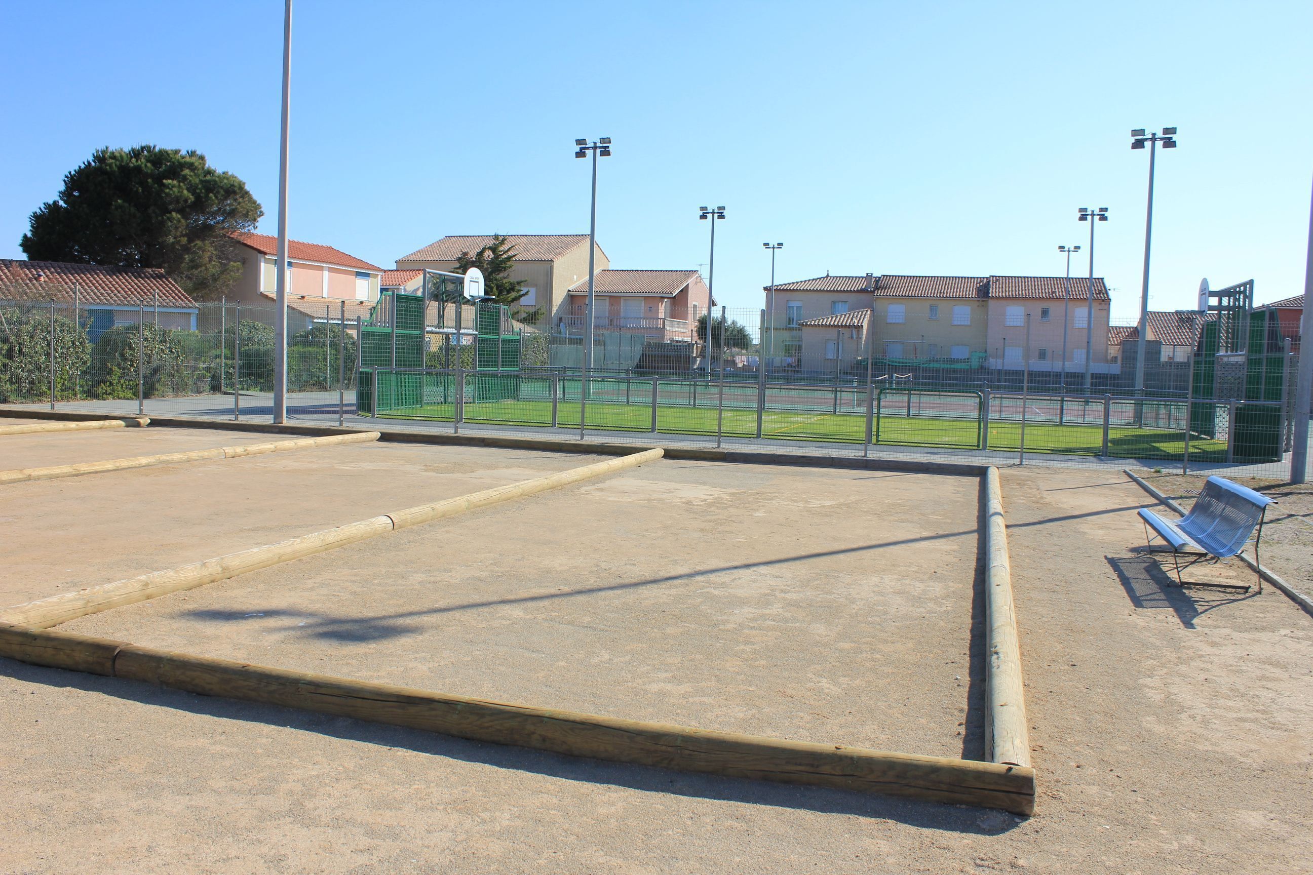 Uma quadra de tênis ao ar livre, com uma cerca verde, um banco e casas ao fundo.