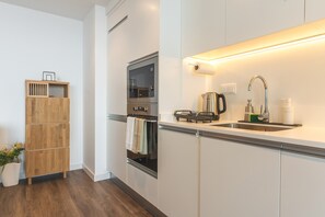 Eine moderne Küche mit Backofen, Wasserkocher und Spüle inklusive Wasserhahn. Ein Holzregal mit Handtüchern und einem Blumentopf.