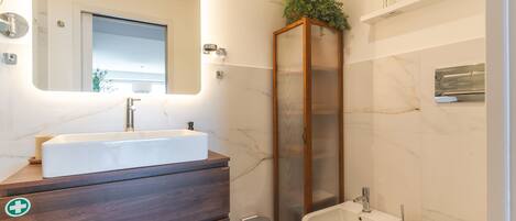 A modern bathroom with a large mirror, a rectangular sink, a bidet, and a tall cabinet with plants.