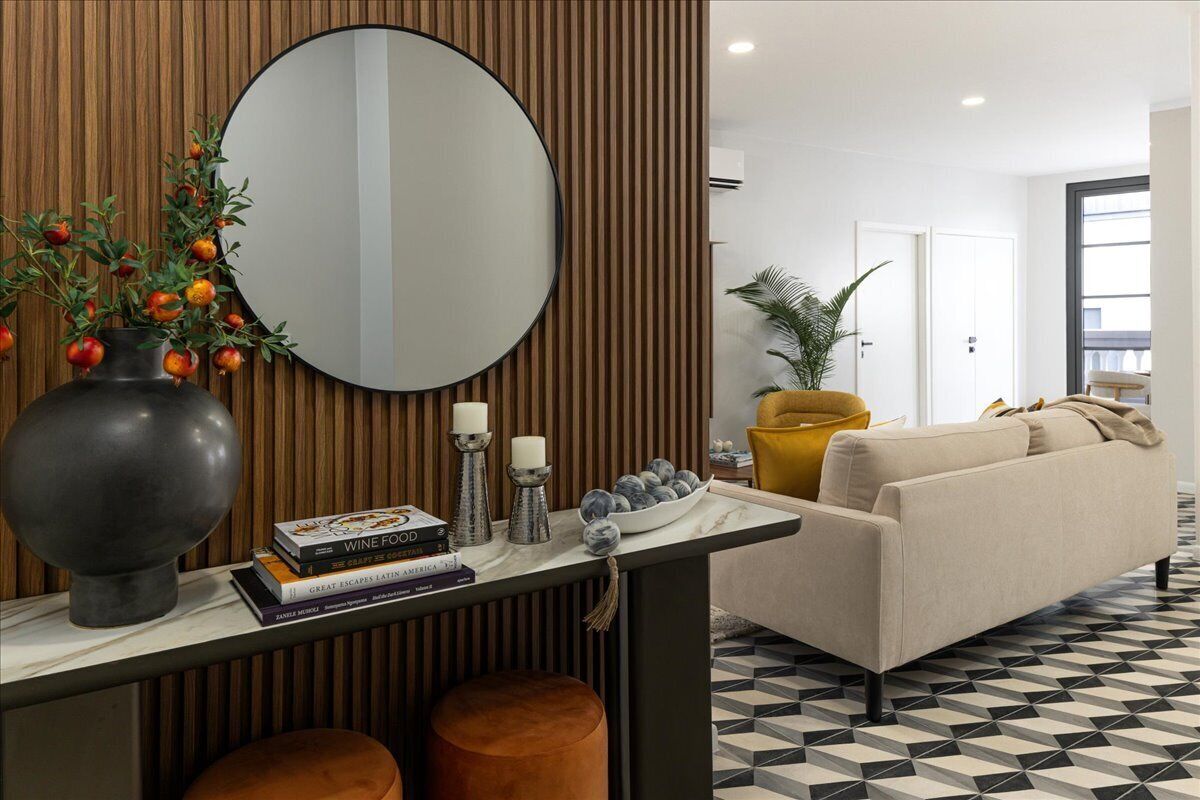 A modern living room with a round mirror, a vase with oranges, a sofa, and a coffee table with books and candles.