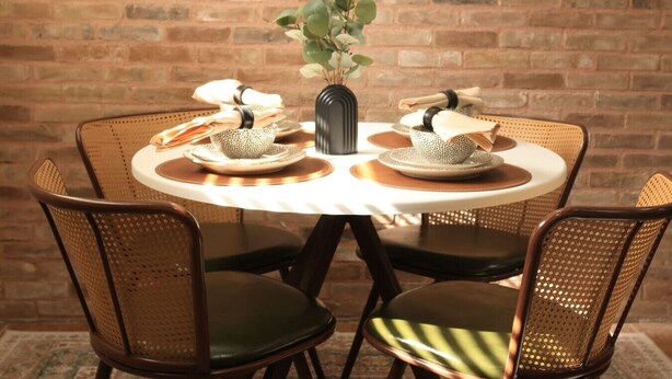 Mesa de jantar redonda com um vaso de plantas, rodeada por cadeiras de vime com almofadas.
