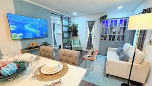 Una sala moderna con un televisor de pantalla plana, un comedor con una mesa preparada para cuatro personas y un sofá.