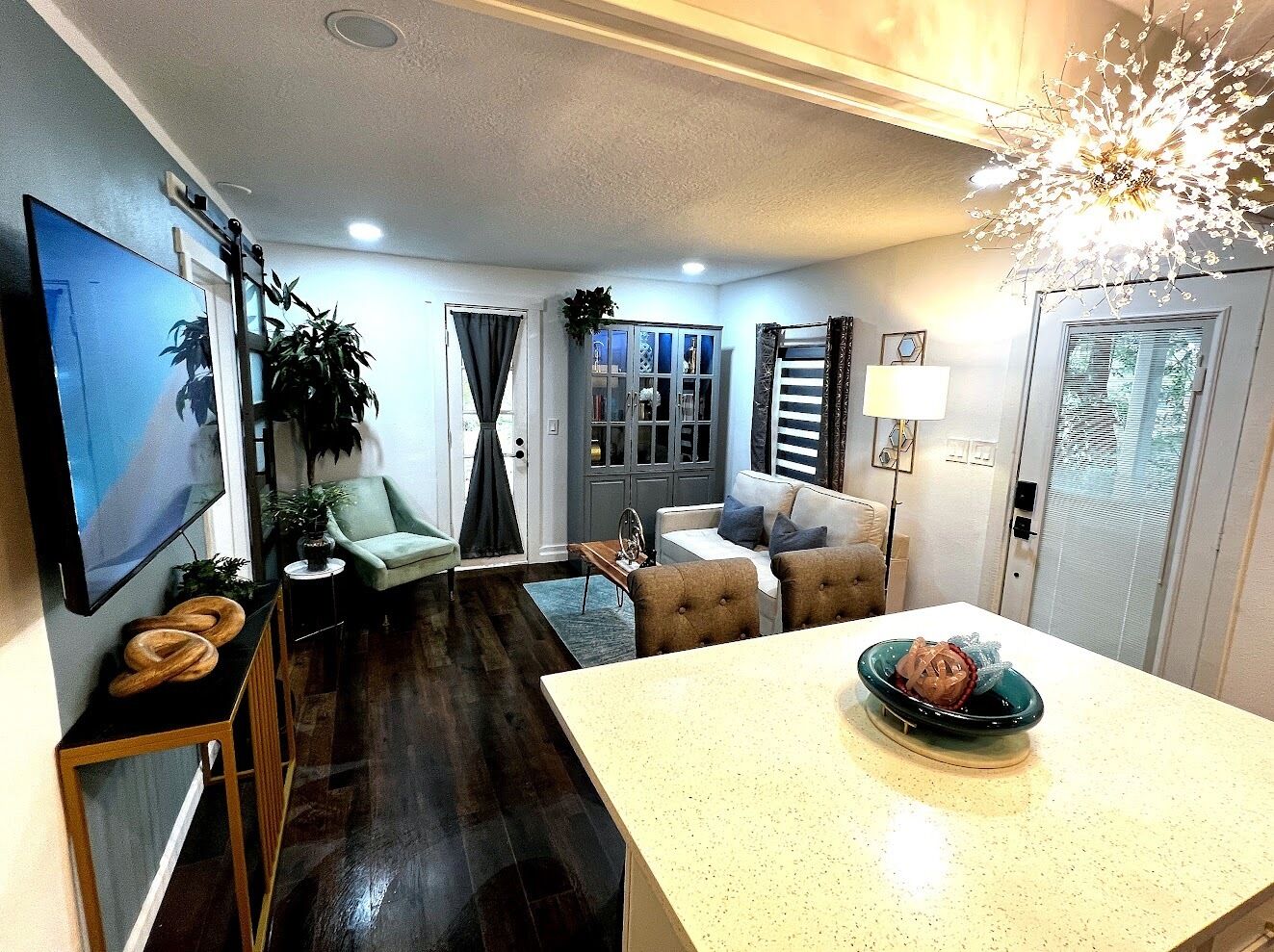 A modern living room with a flat-screen TV, a sofa, a dining table with a decorative bowl, and a kitchen area with cabinets and a refrigerator.
