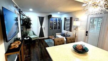 A modern living room with a flat-screen TV, a sofa, a dining table with a decorative bowl, and a kitchen area with cabinets and a refrigerator.