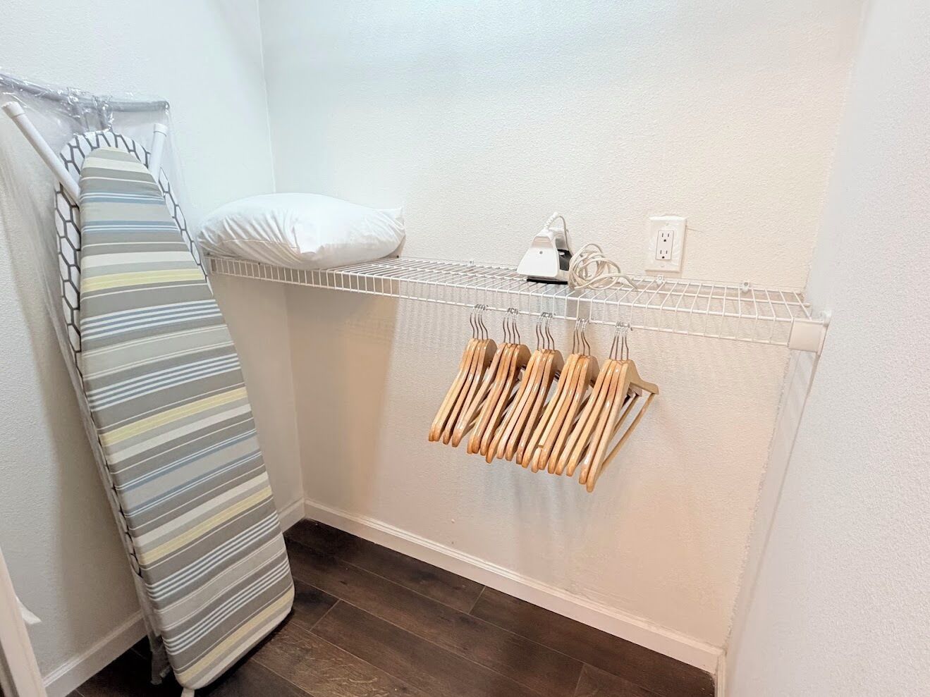A corner of a room with a striped iron board, a white pillow, and a metal drying rack with wooden hangers.