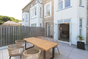 Eine Terrasse mit Holztisch und Korbstühlen, ein Holzzaun und ein Gebäude mit Steinaußenwand und mehreren Fenstern.