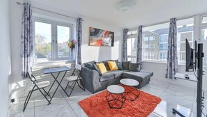 A modern living room with a grey sofa, two white round tables, a red rug, and large windows with curtains. - Cozy 2-bed Escape in Blackheath Smart Studio (London)