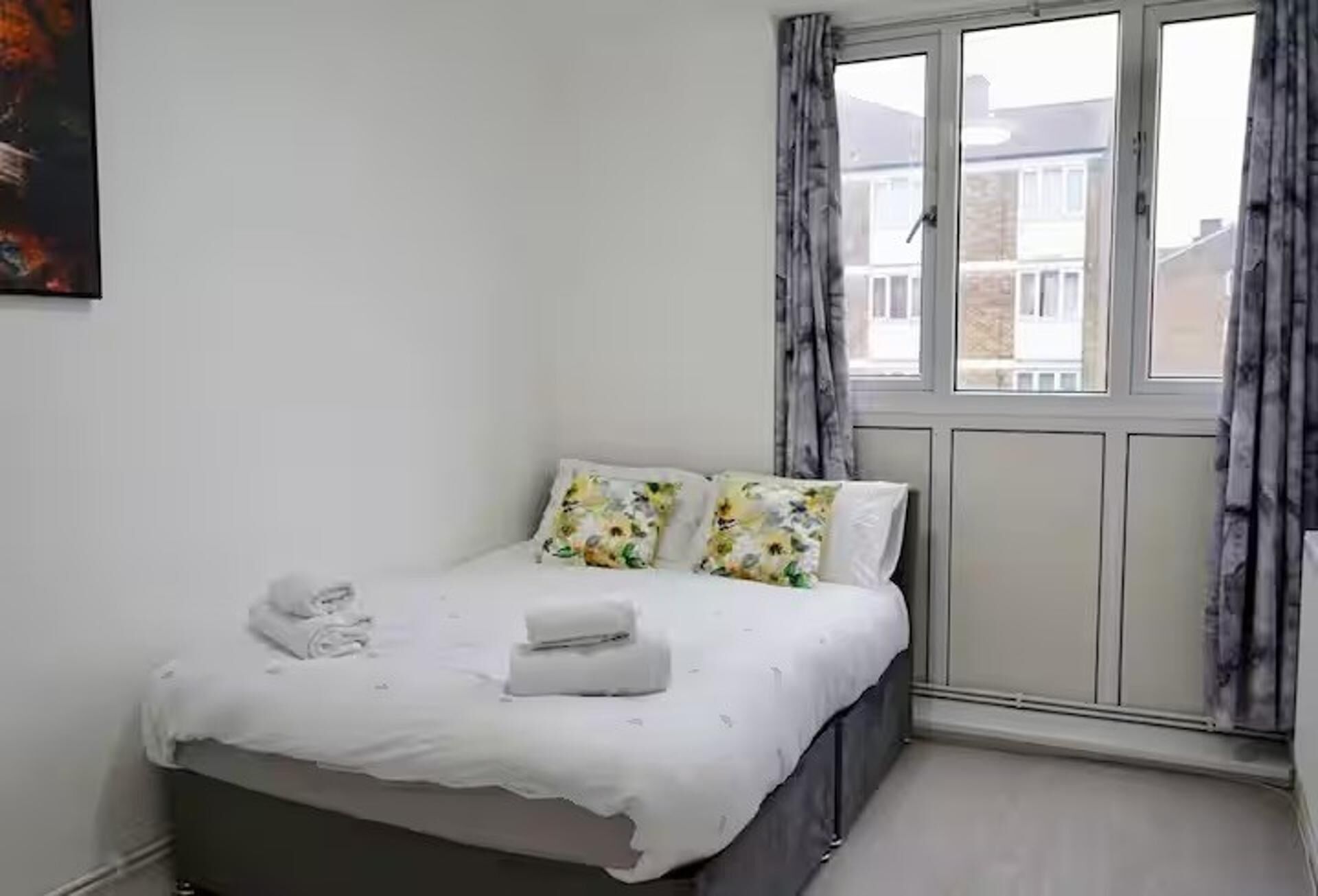 Un dormitorio con cama, almohadas, una ventana con cortinas y un cuadro en la pared.