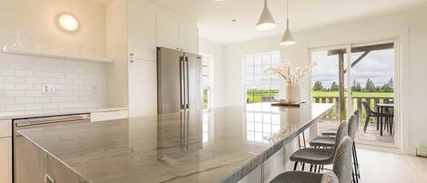 A modern kitchen with a large island, stainless steel appliances, pendant lighting, and a view to the outdoors.