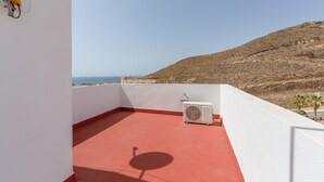A rooftop terrace with a red floor, a white wall, and a view of the sea and hills.
