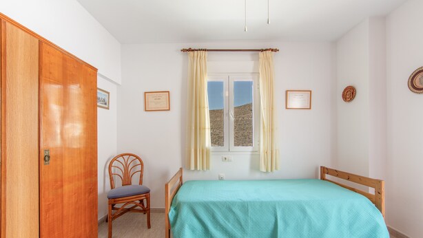 A bedroom with a bed, a wooden wardrobe, a chair, a window with curtains, and two framed certificates on the wall.