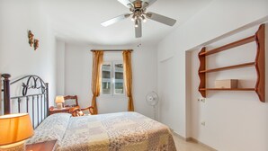 A bedroom with a bed, a nightstand, a lamp, a ceiling fan, a window with curtains, and a wooden shelf. - Holiday Home 'Fmr Fielato 15' with Private Terrace (Carboneras)