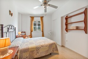 A bedroom with a bed, a nightstand, a lamp, a ceiling fan, a window with curtains, and a wooden shelf.