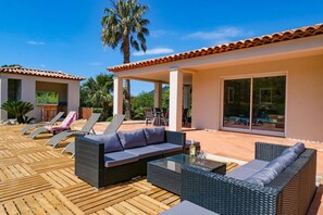An outdoor patio area with wicker furniture, a glass table, and a pool in the background.
