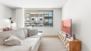 Un salon moderne comprenant un canapé, un téléviseur, une table d’appoint en bois et une fenêtre donnant sur des immeubles.