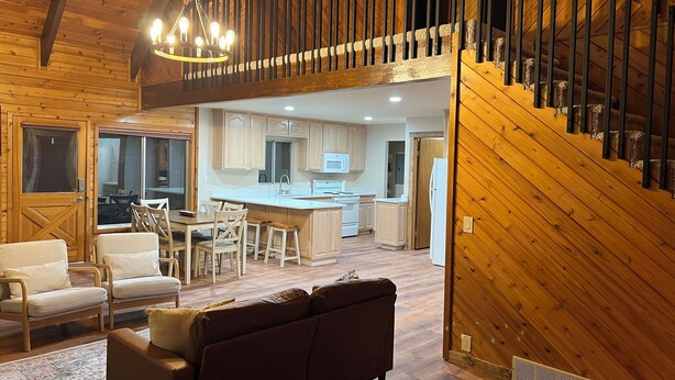A spacious living area with a wooden ceiling, a dining table with chairs, a sofa, and a kitchen with wooden cabinets and a central island.