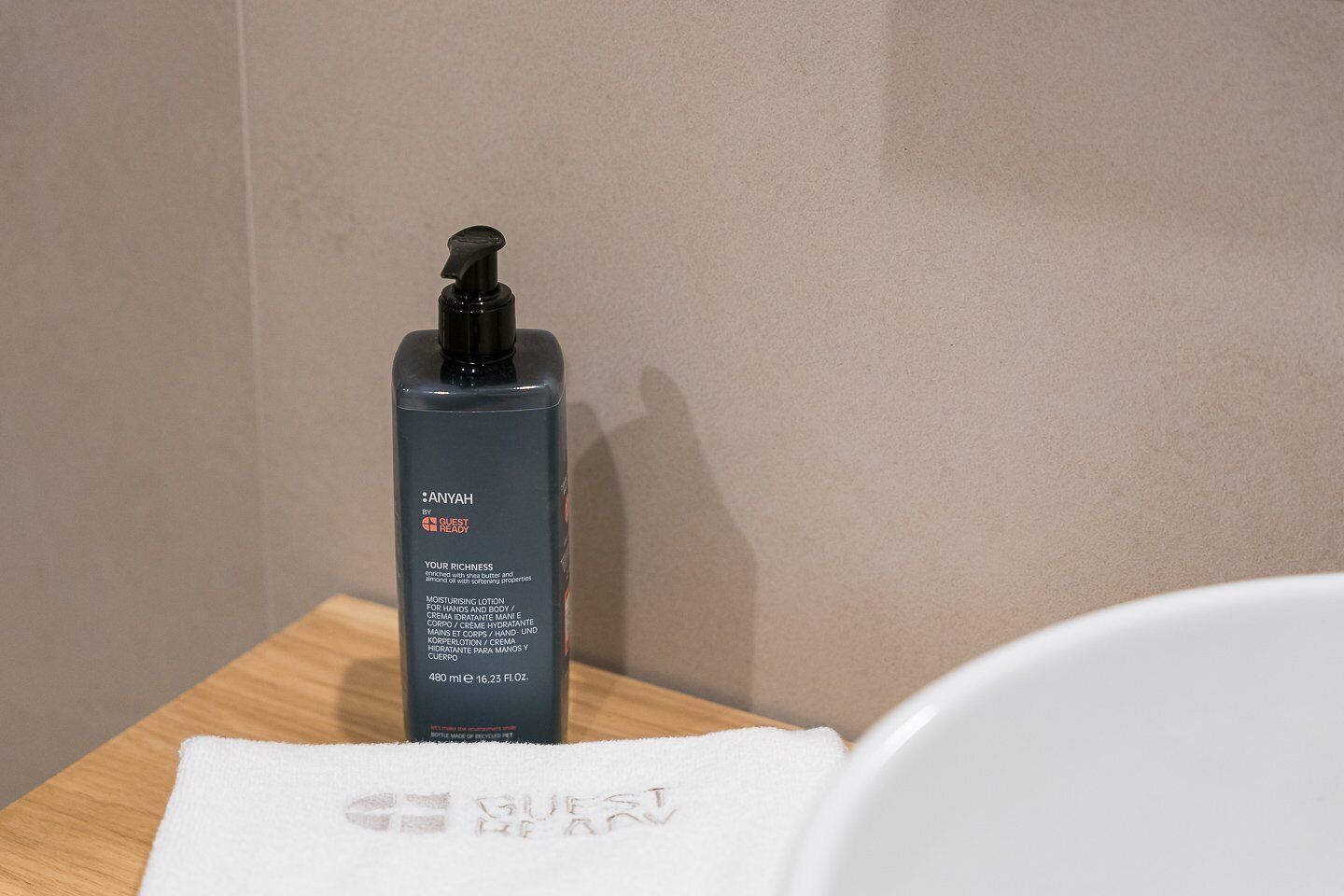 Eine Flasche Anyah Moisturizing Lotion von Guest Ready auf einer Holzoberfläche neben einem weißen Handtuch mit dem aufgedruckten Schriftzug 'GUEST READY'.
