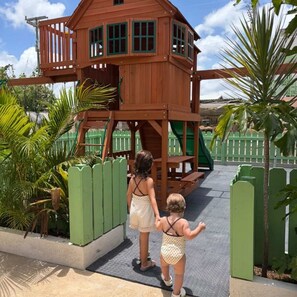 A wooden play structure with a slide, surrounded by green fencing and palm trees. - Sunfish 1-bedroom ocean view apartment in Sand Street, Speightstown Apt 2 (Speightstown)