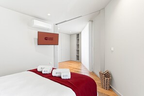 Una habitación de hotel con una cama, una manta roja, un televisor montado en la pared y un pasillo que conduce a una escalera.