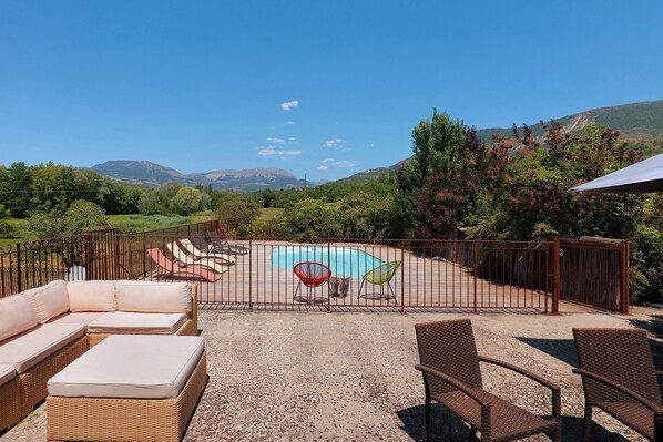 A pool area with a view of mountains, surrounded by a fence, and furnished with lounge chairs and a sofa.