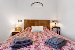 A bedroom with a wooden headboard, a patterned bedspread, two bedside tables with lamps, and a wall-mounted light fixture.