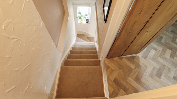 A staircase with carpeted steps leading to a landing, a wooden door, and a herringbone-patterned floor. - Pax Cottage (Beccles)
