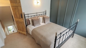 A bedroom with a bed, a wooden wardrobe, a wall-mounted light fixture, and a window with a plant. - Pax Cottage (Beccles)