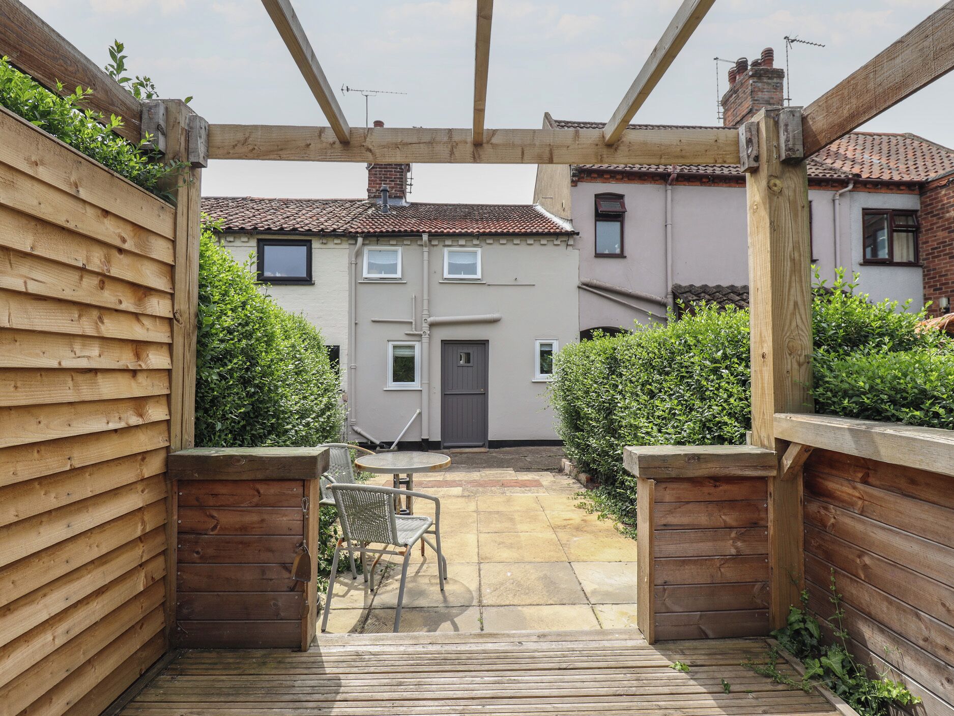 Eine hölzerne Pergola mit Tisch und Stühlen, die zu einem Haus mit Ziegeldach und einer Tür führt.