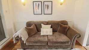A brown sofa with a white throw blanket and pillows, two framed feather pictures on the wall, and a white radiator below. - Pax Cottage (Beccles)