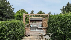 A wooden pergola with a cushioned seating area and a small table and chair set. - Pax Cottage (Beccles)