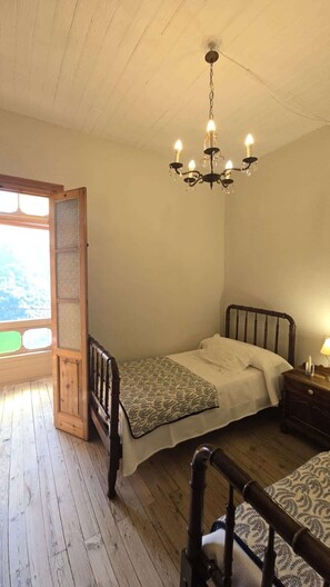 A bedroom with a wooden floor, a bed with a patterned blanket, a wooden headboard, a nightstand with a lamp, and a window with a view.