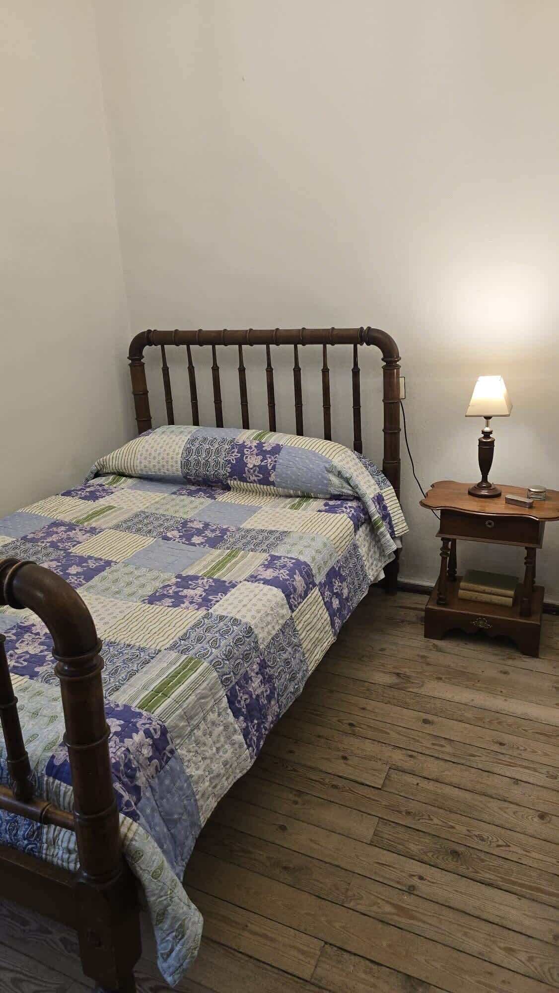 A single bed with a wooden headboard, a quilt with floral patterns, a small wooden nightstand with a lamp, and a wooden floor.